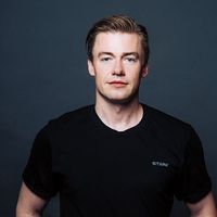 Mann mit kurzem Haar trägt ein schwarzes T-Shirt mit dem Schriftzug 'STARK' vor dunklem Hintergrund