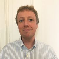 Mann mit kurz geschnittenem, lockigem Haar trägt ein hellblau-weiß gestreiftes Hemd mit Knopfleiste vor einer hellen Wand